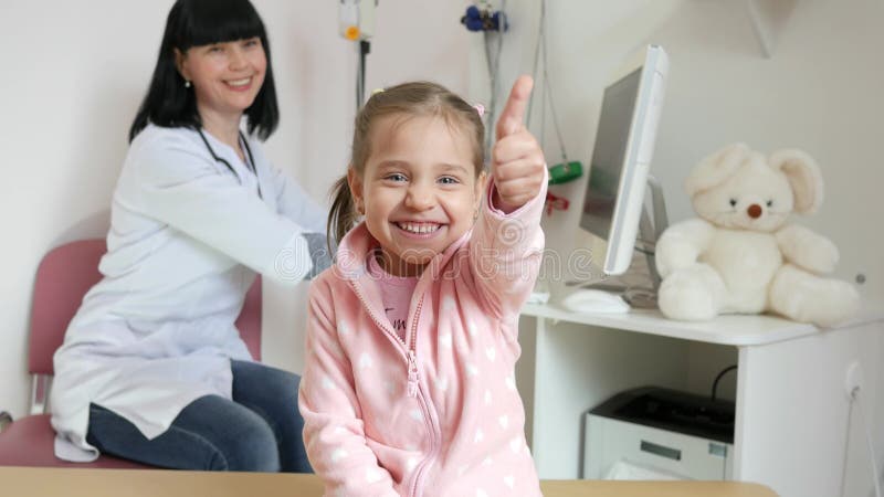 O doutor móvel, menina bonito mostra o sinal da aprovação, retrato da criança na clínica médica, tratamento paciente em linha, do