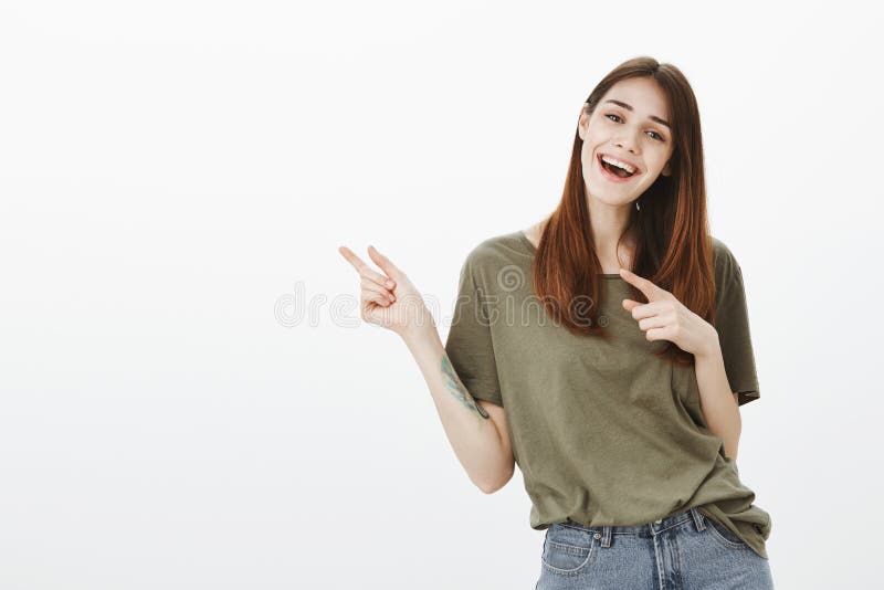 O estúdio disparou da mulher brincalhão feliz do partido com cabelo marrom, sorrindo amplamente e felizmente ao apontar com indic fotografia de stock O estúdio disparou da mulher brincalhão feliz do partido com cabelo marrom, sorrindo amplamente e felizmente ao apontar com indic fotografia de stock