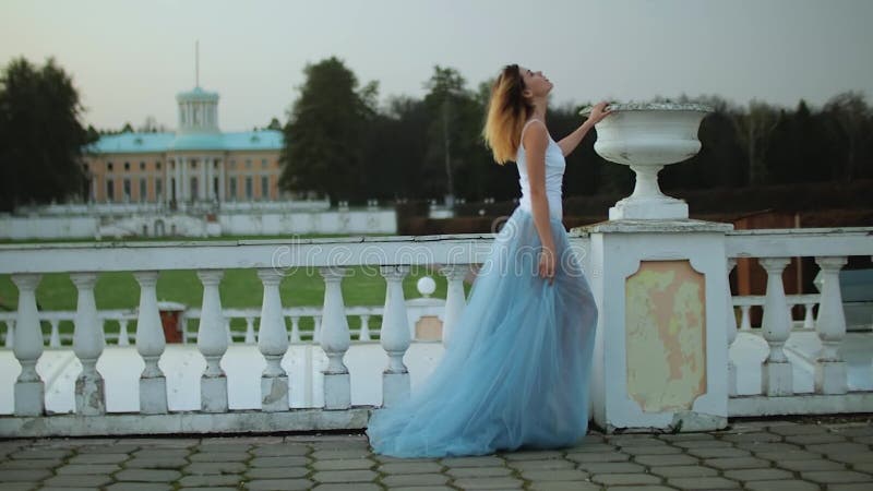 O fată atrăgătoare și slabă în rochie albă și albastră stă lângă balustrade de piatră albă, cu ghiveci înflorit în timp ce se prez stock footage