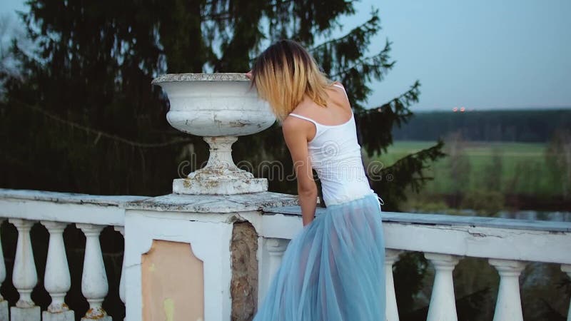 O fată atrăgătoare și slabă în rochie albă și albastră stă lângă balustrade de piatră albă, cu ghiveci înfloriți pe ea și arată stock footage