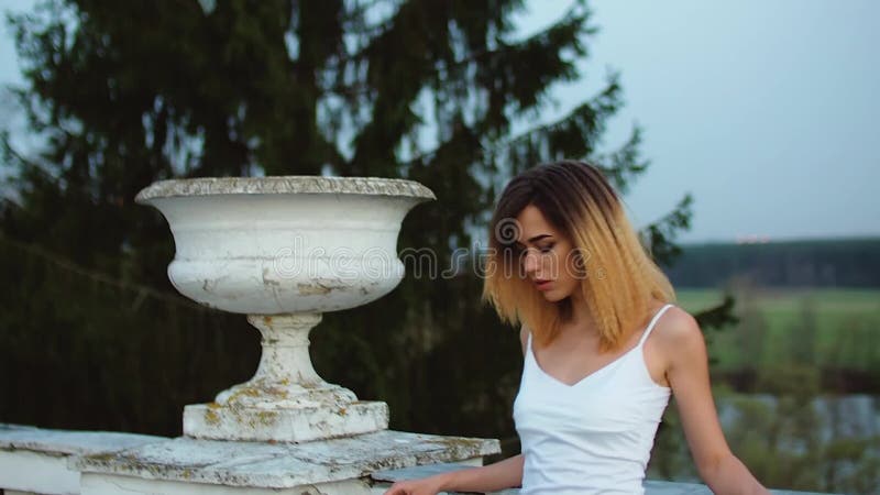 O fată atrăgătoare și slabă în rochie albă și albastră stă lângă balustrade de piatră albă, cu ghiveci înfloriți pe ea și arată stock footage