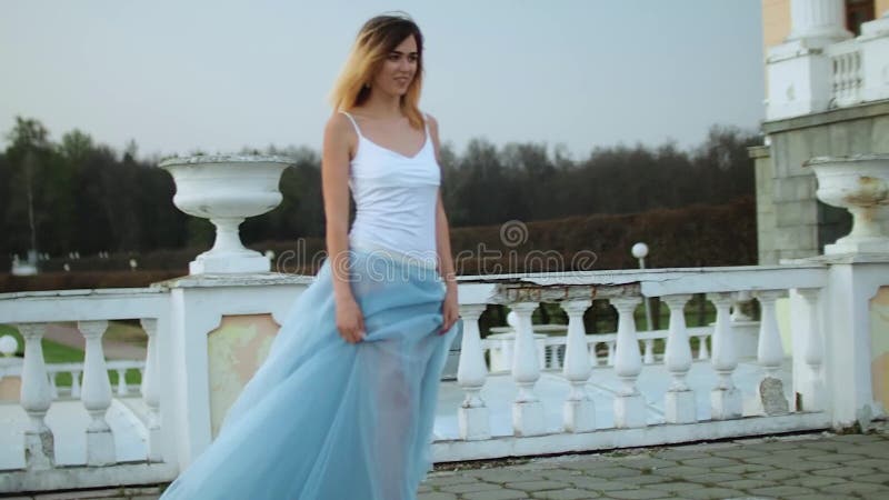 O fată atrăgătoare și slabă în rochie albă și albastră stă lângă balustrade de piatră albă cu un ghiveci cu flori care se ridică ș stock video footage
