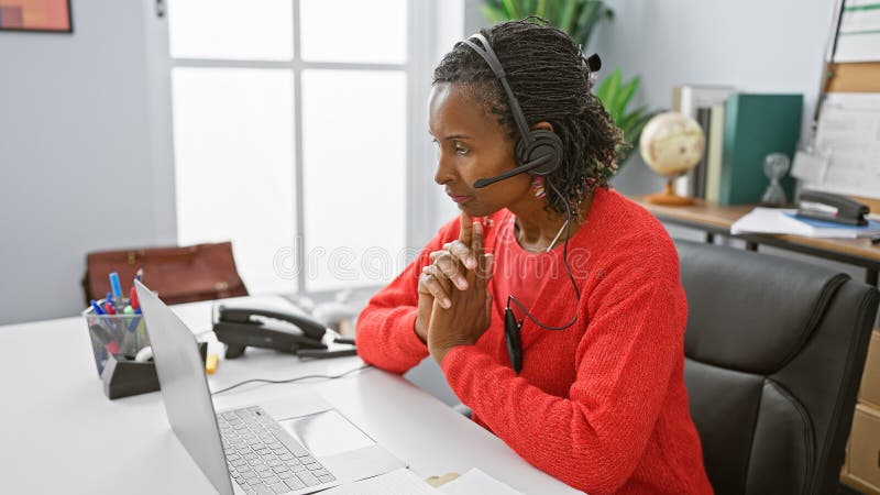 O femeie americană africană care utilizează un laptop și căști într-un birou modern, orientat și profesionist royalty free stock images