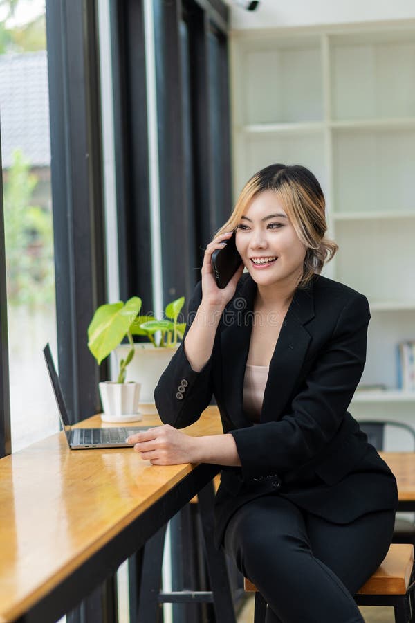 O femeie asiatică frumoasă care vorbește pe telefonul mobil și se joacă cu un notebook care zâmbește în timp ce stă la birou. part royalty free stock photography