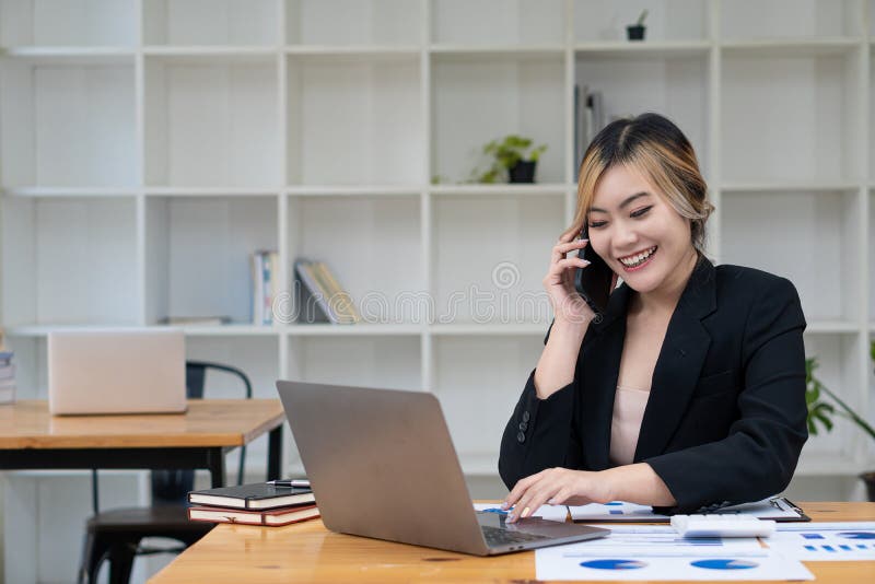 O femeie asiatică vorbește pe un telefon mobil sau smartphone în timp ce lucrează pe un calculator la biroul său. royalty free stock photo