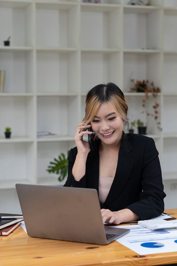 O femeie asiatică vorbește pe un telefon mobil sau smartphone în timp ce lucrează pe un calculator la biroul său. stock image