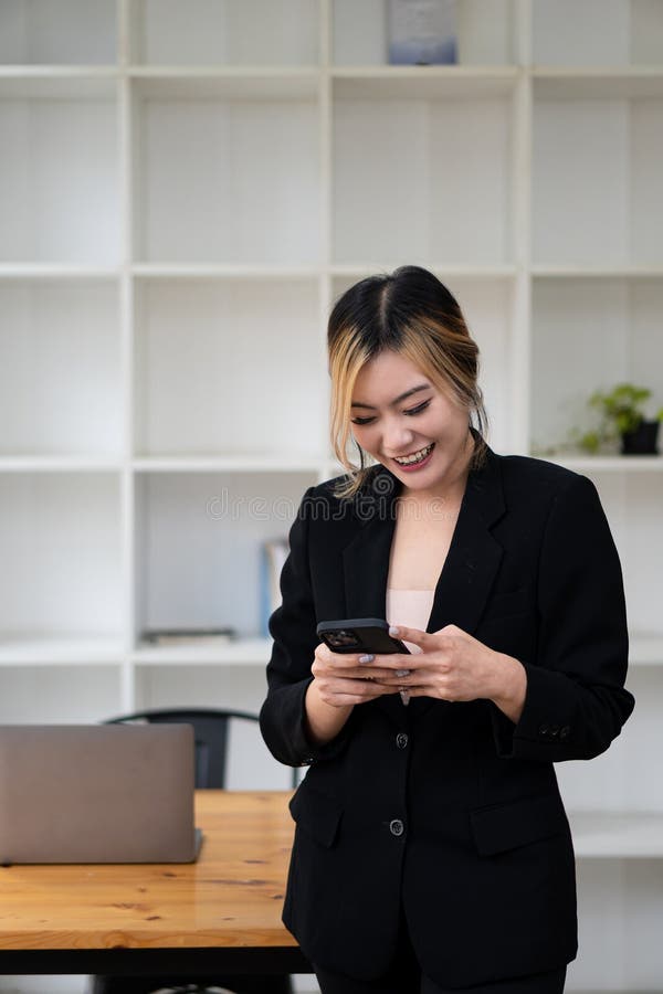 O femeie asiatică vorbește pe un telefon mobil sau smartphone în timp ce lucrează pe un calculator la biroul său. stock image