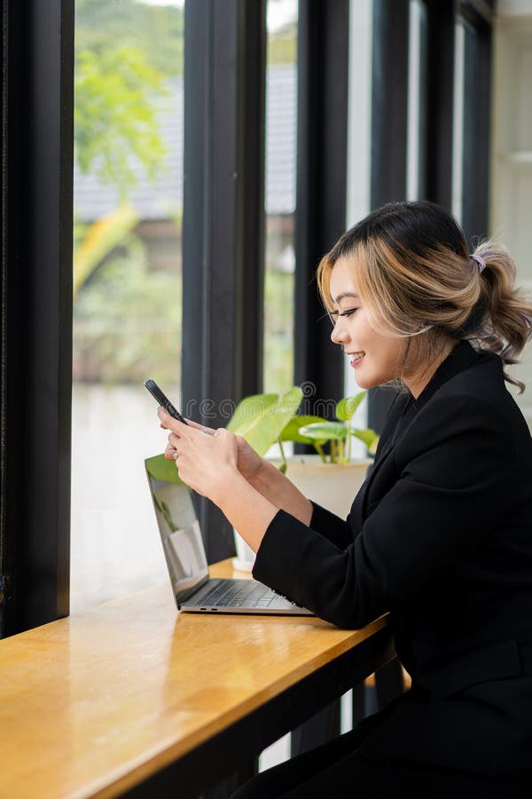 O femeie asiatică vorbește pe un telefon mobil sau smartphone în timp ce lucrează pe un calculator la biroul său. stock images