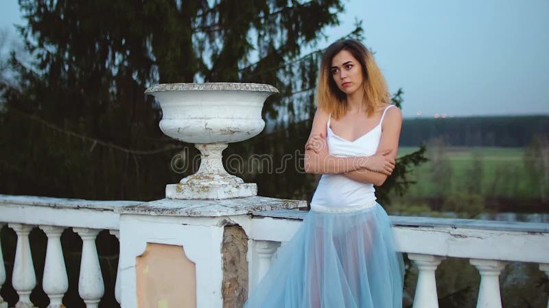 O fetiță atrăgătoare, în rochie albă și albastră, se află lângă balustrade de piatră albă, cu ghiveci cu brațe încrucișate și stock video