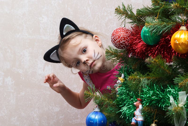 O gato pintou a menina que espreita para fora da árvore de Natal de trás imagem de stock royalty free O gato pintou a menina que espreita para fora da árvore de Natal de trás imagem de stock royalty free