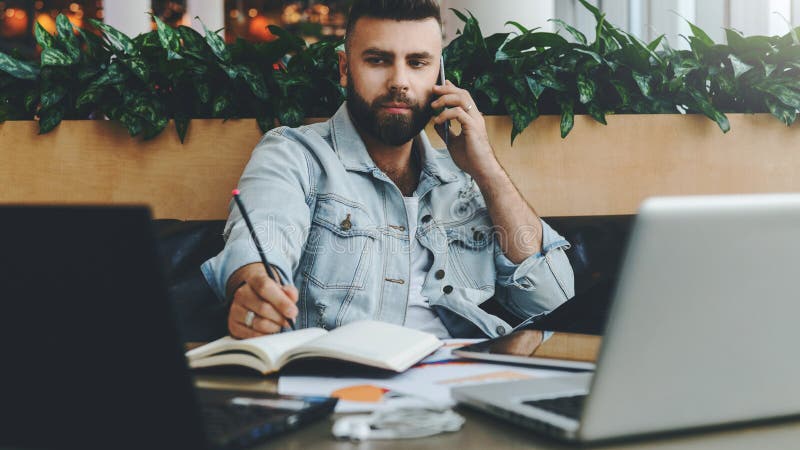 O homem alegre farpado novo senta-se na tabela na frente dos portáteis, falando no telefone celular ao fazer anotações no caderno fotografia de stock royalty free O homem alegre farpado novo senta-se na tabela na frente dos portáteis, falando no telefone celular ao fazer anotações no caderno fotografia de stock royalty free