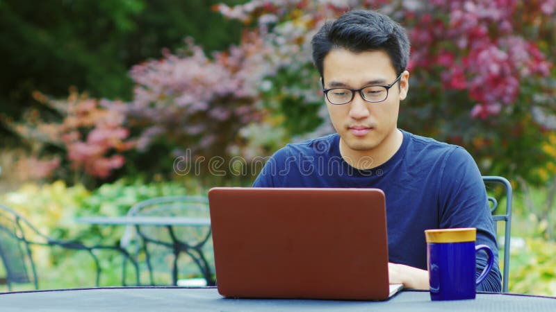 O homem asiático que veste vidros trabalha com um portátil Assento no terraço do verão nos jardins ou nos cafés