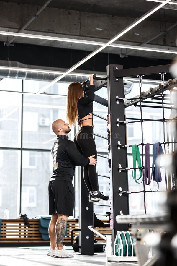 O homem atlético ajuda a menina delgada nova vestida na roupa preta dos tipos para fazer levanta na barra no gym imagens de stock royalty free O homem atlético ajuda a menina delgada nova vestida na roupa preta dos tipos para fazer levanta na barra no gym imagens de stock royalty free