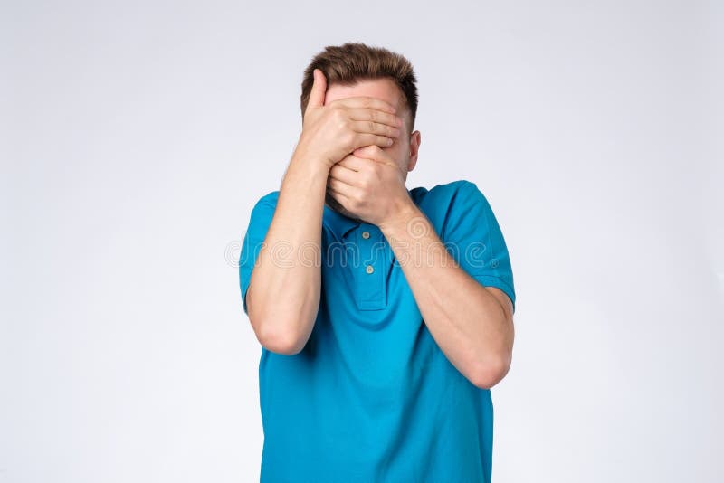 O homem caucasiano novo na camisa azul fecha a cara com as mãos que tentam ficar o anônimo fotos de stock royalty free O homem caucasiano novo na camisa azul fecha a cara com as mãos que tentam ficar o anônimo fotos de stock royalty free