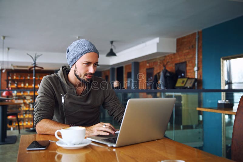 O homem considerável novo do moderno com barba está usando o portátil no bar imagem de stock royalty free O homem considerável novo do moderno com barba está usando o portátil no bar imagem de stock royalty free