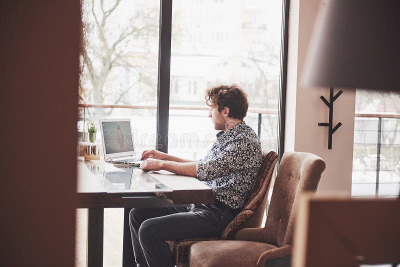 O homem considerável novo que senta-se no escritório com xícara de café e que trabalha no projeto conectou com as tecnologias mod foto de stock royalty free O homem considerável novo que senta-se no escritório com xícara de café e que trabalha no projeto conectou com as tecnologias mod foto de stock royalty free