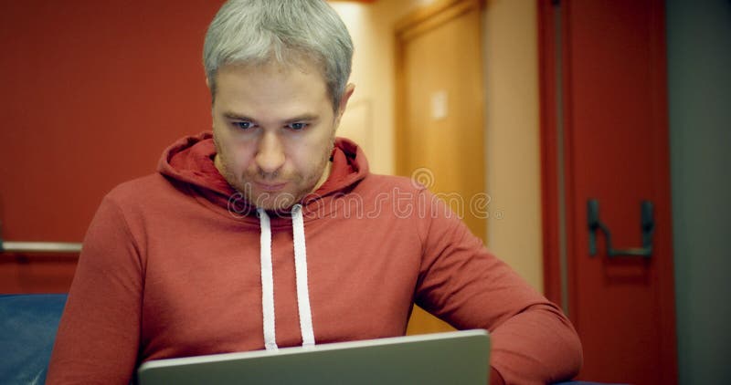 O homem de cabelo cinzento esperto novo na roupa ocasional usa seu portátil na entrada foto de stock O homem de cabelo cinzento esperto novo na roupa ocasional usa seu portátil na entrada foto de stock