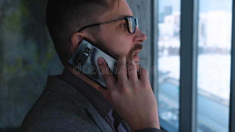 O homem de negócios bem sucedido novo com uma barba faz chamar um smartphone em um fundo da janela com uma vista da baixa