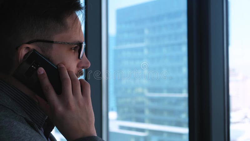 O homem de negócios bem sucedido novo com uma barba faz chamar um smartphone em um fundo da janela com uma vista da baixa