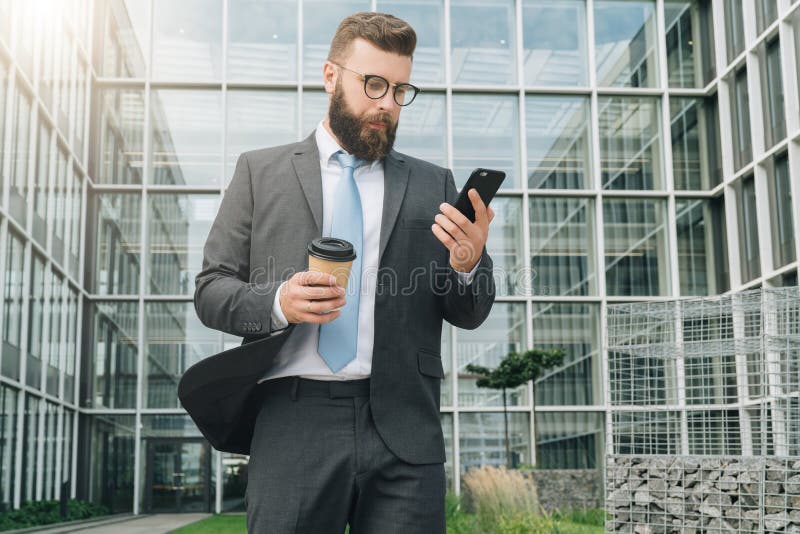 O homem de negócios novo nos monóculos, no terno e no laço está estando exterior, está usando o smartphone e está bebendo o café imagem de stock royalty free O homem de negócios novo nos monóculos, no terno e no laço está estando exterior, está usando o smartphone e está bebendo o café imagem de stock royalty free