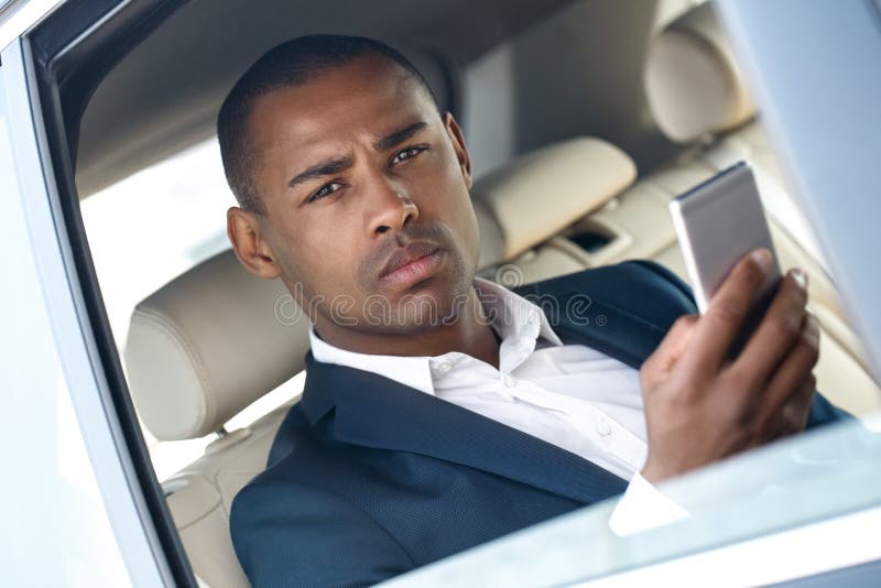 O homem de negócios novo que senta-se na janela de carro abriu o smartphone da consultação que olha o close-up pensativo da câmer fotografia de stock royalty free O homem de negócios novo que senta-se na janela de carro abriu o smartphone da consultação que olha o close-up pensativo da câmer fotografia de stock royalty free