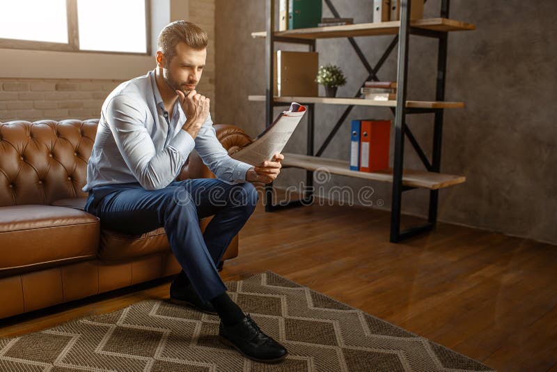 O homem de negócios pensativo considerável novo senta-se no sofá e no jornal lido em seu próprio escritório Levanta Retrato 'sexy fotos de stock royalty free O homem de negócios pensativo considerável novo senta-se no sofá e no jornal lido em seu próprio escritório Levanta Retrato 'sexy fotos de stock royalty free