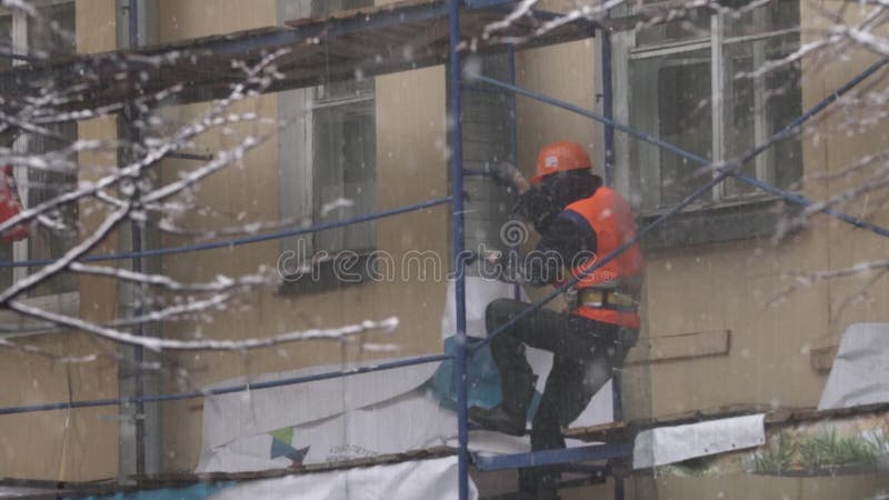 O homem do trabalhador no uniforme e no capacete vai abaixo das escadas do andaime da construção