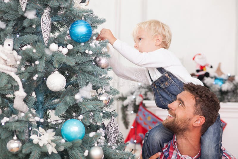 Cheerful father and son are preparing Christmas tree for celebration. The boy is hanging sphere with concentration. His parent is holding him and looking at child happily. He is smiling. Cheerful father and son are preparing Christmas tree for celebration. The boy is hanging sphere with concentration. His parent is holding him and looking at child happily. He is smiling