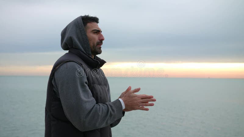 O homem está aquecendo suas mãos respirando no beira-mar frio da manhã Nascer do sol da manhã sobre o mar