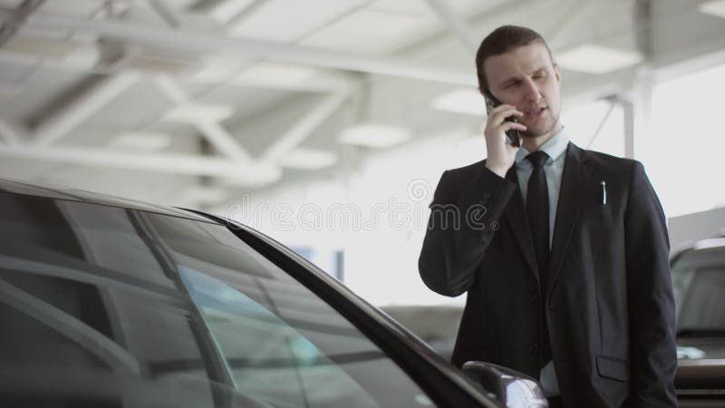 O homem está escolhendo um carro ou ele é um vendedor de carros. O jovem fala por telefone em pé no salão de automóvel.