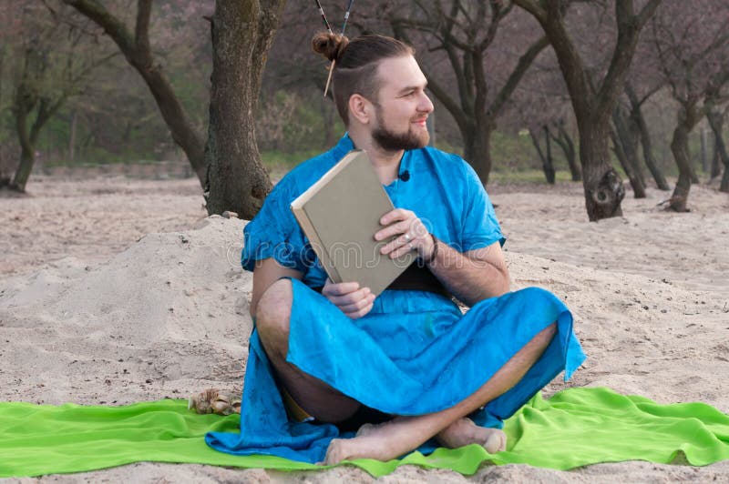 O homem farpado considerável com compõe, bolo na cabeça no assento azul do quimono, guardando o livro fechado foto de stock O homem farpado considerável com compõe, bolo na cabeça no assento azul do quimono, guardando o livro fechado foto de stock