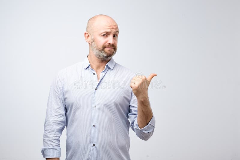 O homem farpado considerável na roupa ocasional está apontando o polegar do awaywith que olha a câmera no fundo cinzento fotografia de stock O homem farpado considerável na roupa ocasional está apontando o polegar do awaywith que olha a câmera no fundo cinzento fotografia de stock