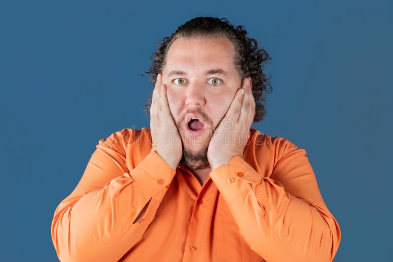 O homem gordo na camisa alaranjada guarda suas mãos sobre sua cara É surpreendido muito imagens de stock O homem gordo na camisa alaranjada guarda suas mãos sobre sua cara É surpreendido muito imagens de stock