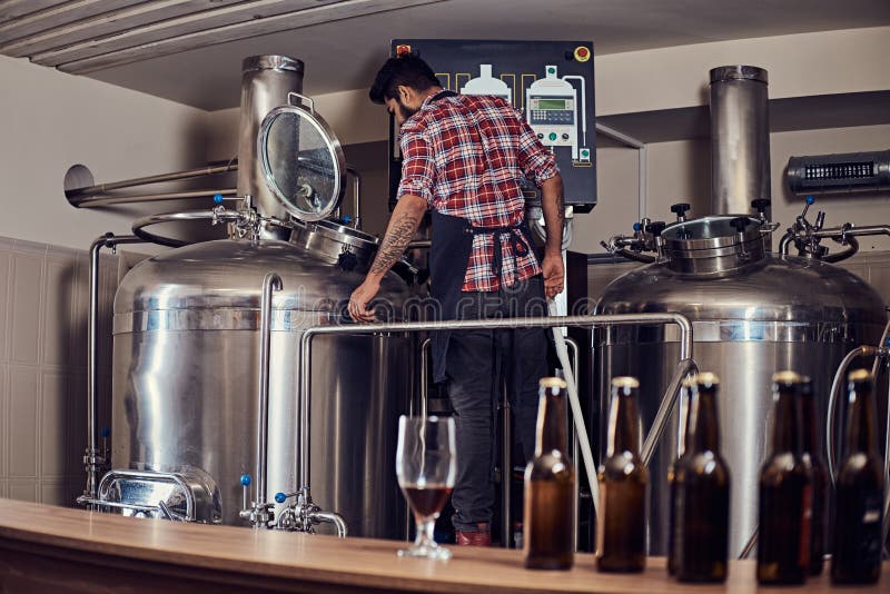 O homem indiano farpado completo à moda em uma camisa e em um avental do velo controla o processo da fabricação de cerveja, estan imagem de stock royalty free O homem indiano farpado completo à moda em uma camisa e em um avental do velo controla o processo da fabricação de cerveja, estan imagem de stock royalty free