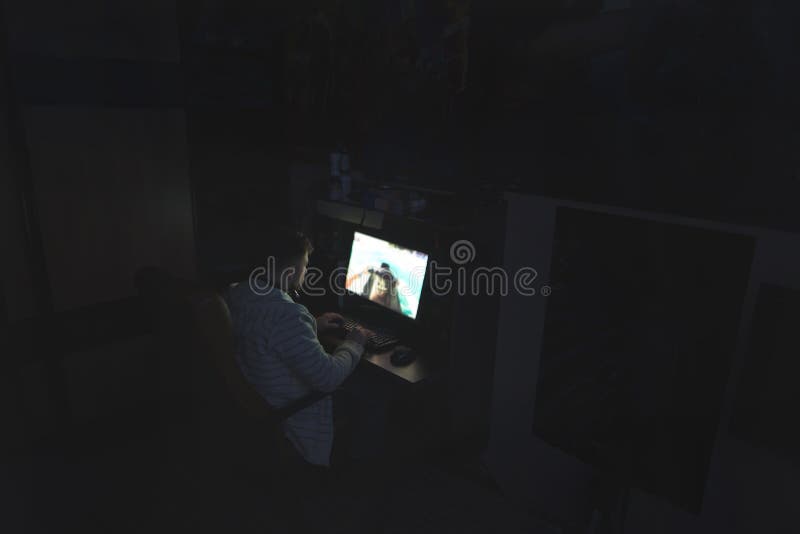 O homem joga jogos em casa em uma sala escura Jogos noturnos no computador O Gamer joga jogos de computador na noite imagem de stock royalty free O homem joga jogos em casa em uma sala escura Jogos noturnos no computador O Gamer joga jogos de computador na noite imagem de stock royalty free