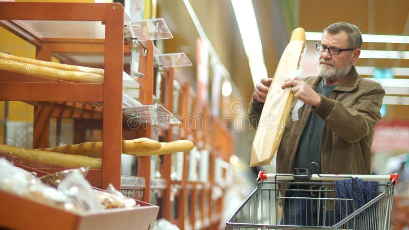 O homem maduro com uma barba cinzenta e os passeios dos vidros com um trole através de um supermercado, compram um baguette fresc