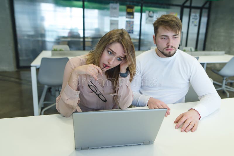 O homem novo e a menina tristes estão sentando-se em uma mesa perto de um portátil no escritório e são tristes Falha no trabalho foto de stock royalty free O homem novo e a menina tristes estão sentando-se em uma mesa perto de um portátil no escritório e são tristes Falha no trabalho foto de stock royalty free