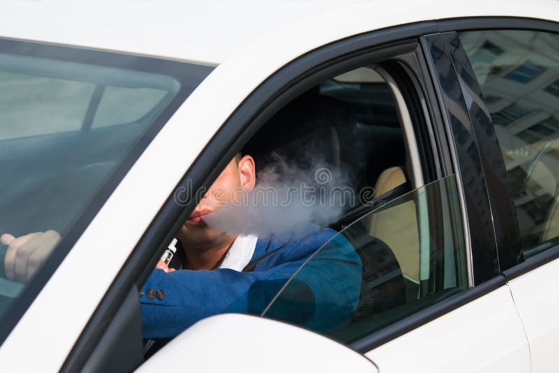 O homem novo em um casaco azul fuma um cigarro eletrônico ao conduzir um carro foto de stock O homem novo em um casaco azul fuma um cigarro eletrônico ao conduzir um carro foto de stock
