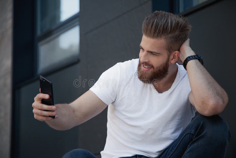 O homem novo farpado considerável está fazendo o selfie e está sorrindo ele está na camisa branca de t imagem de stock O homem novo farpado considerável está fazendo o selfie e está sorrindo ele está na camisa branca de t imagem de stock