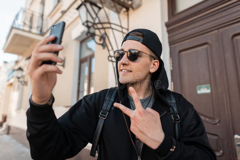 O homem novo feliz atrativo do moderno com um sorriso bonito na roupa preta elegante em um tampão nos óculos de sol faz um selfie imagens de stock royalty free O homem novo feliz atrativo do moderno com um sorriso bonito na roupa preta elegante em um tampão nos óculos de sol faz um selfie imagens de stock royalty free