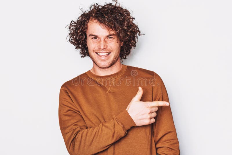 O homem novo feliz com cabelo encaracolado veste o pulôver indica um lado com o dedo dianteiro, sorrindo e demonstra algo na plac fotos de stock O homem novo feliz com cabelo encaracolado veste o pulôver indica um lado com o dedo dianteiro, sorrindo e demonstra algo na plac fotos de stock