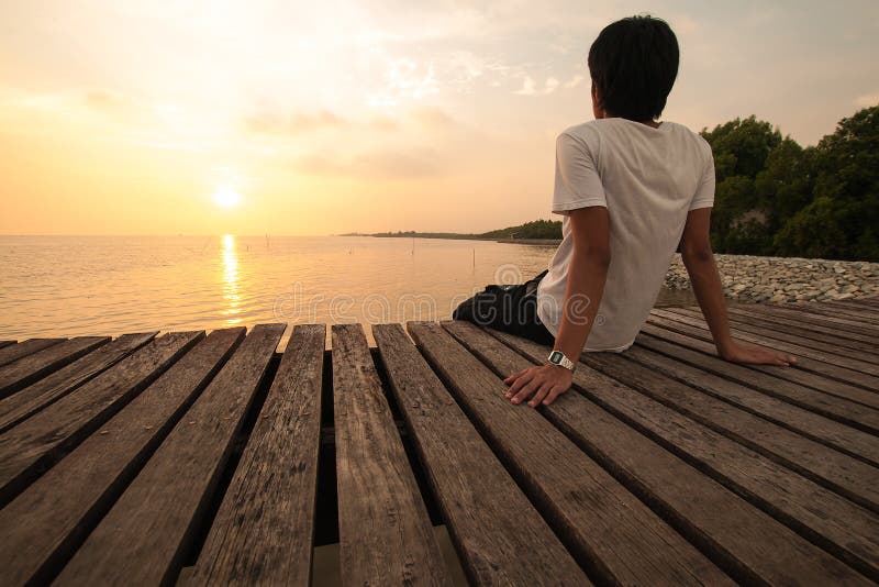 O homem novo relaxa a situação em olhares do cais para a frente com por do sol imagem de stock royalty free O homem novo relaxa a situação em olhares do cais para a frente com por do sol imagem de stock royalty free