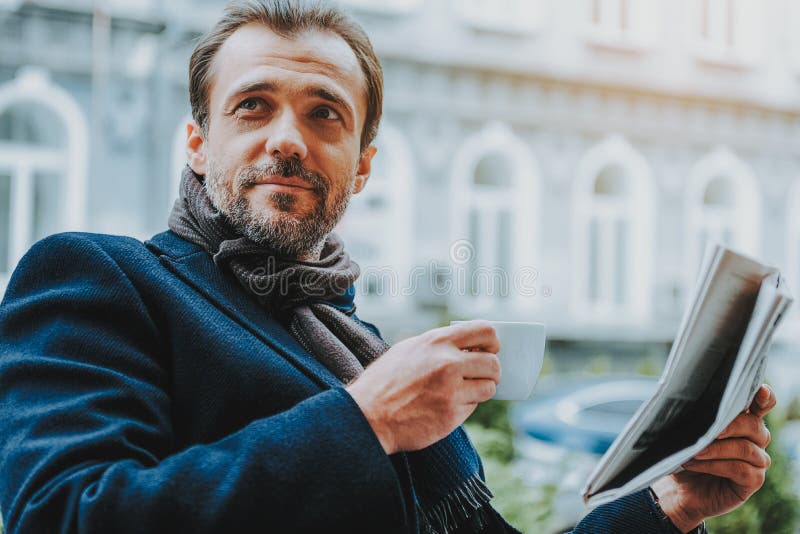 O homem positivo está tendo a bebida quente e está lendo o papel fora imagem de stock O homem positivo está tendo a bebida quente e está lendo o papel fora imagem de stock