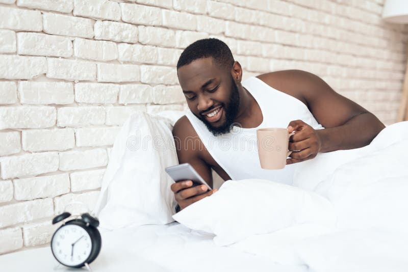 O homem preto, despertado bebe o café na cama que consulta foto de stock O homem preto, despertado bebe o café na cama que consulta foto de stock