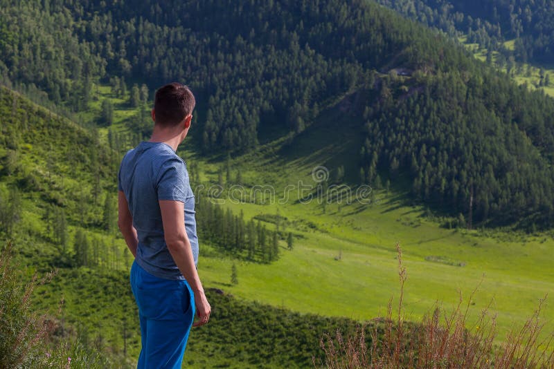O indivíduo em um t-shirt verde está estando na borda do penhasco na parte traseira dele é uma vista colorida das montanhas e de foto de stock royalty free O indivíduo em um t-shirt verde está estando na borda do penhasco na parte traseira dele é uma vista colorida das montanhas e de foto de stock royalty free