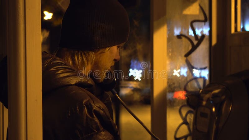 O indivíduo novo é de chamada e falador na cabine de telefone na cidade da noite, tempo frio