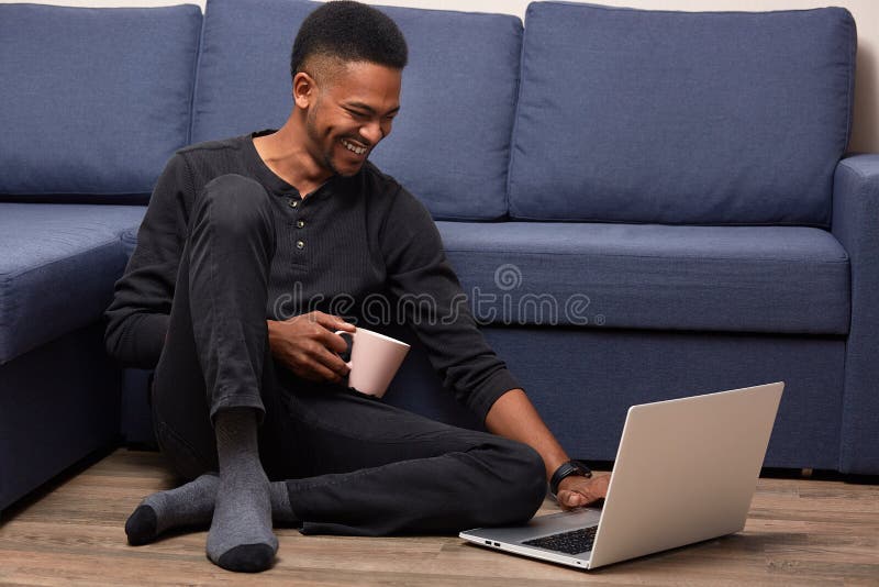 O indoorm horizontal disparou do homem descascado escuro que levanta no assoalho perto do sofá azul, olha o vídeo engraçado no la foto de stock royalty free O indoorm horizontal disparou do homem descascado escuro que levanta no assoalho perto do sofá azul, olha o vídeo engraçado no la foto de stock royalty free