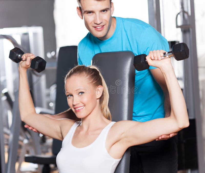 O instrutor que ajuda uma mulher elabora com dumbbells fotografia de stock O instrutor que ajuda uma mulher elabora com dumbbells fotografia de stock