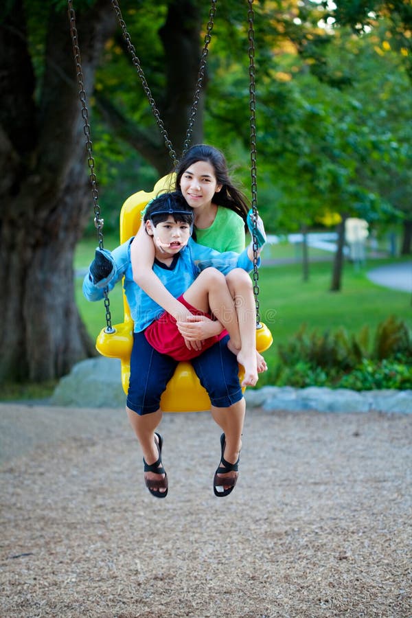 Big sister holding disabled brother on special needs swing at playground in park. Child has cerebral palsy. Big sister holding disabled brother on special needs swing at playground in park. Child has cerebral palsy.