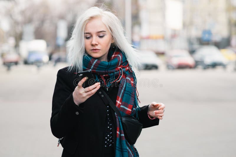 O louro bonito vestiu-se na roupa morna que anda abaixo da rua e que olha o telefone celular Retrato ao ar livre fotos de stock royalty free O louro bonito vestiu-se na roupa morna que anda abaixo da rua e que olha o telefone celular Retrato ao ar livre fotos de stock royalty free